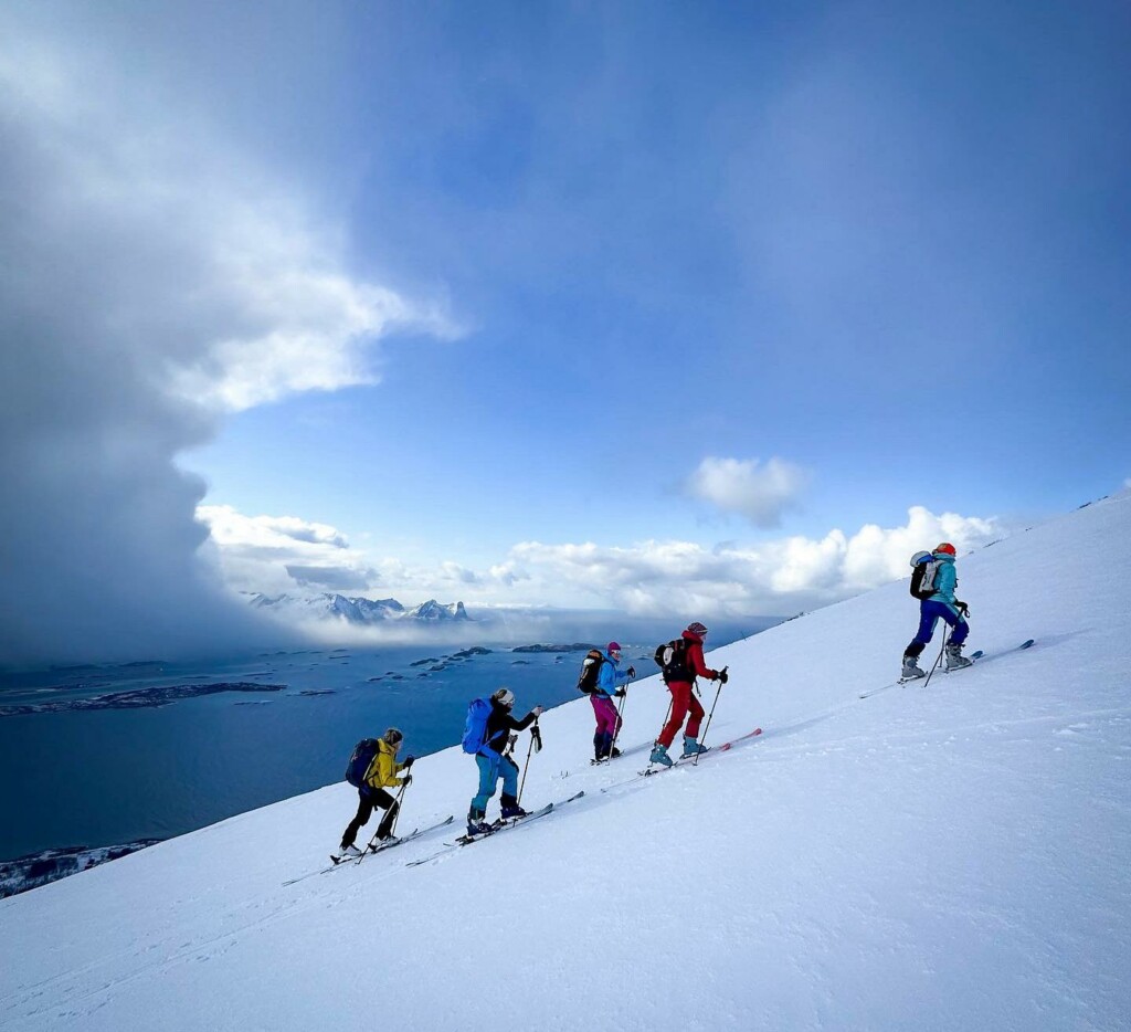 Five ski tourers skinning up a snow slope above Lyngenfjord, with cloud over the far range